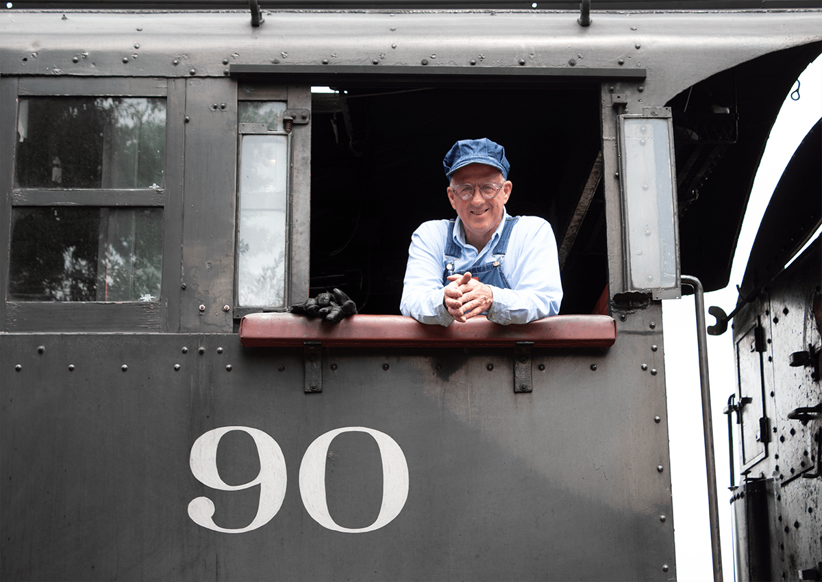 A male conductor smiling down from the window of a steam locomotive.