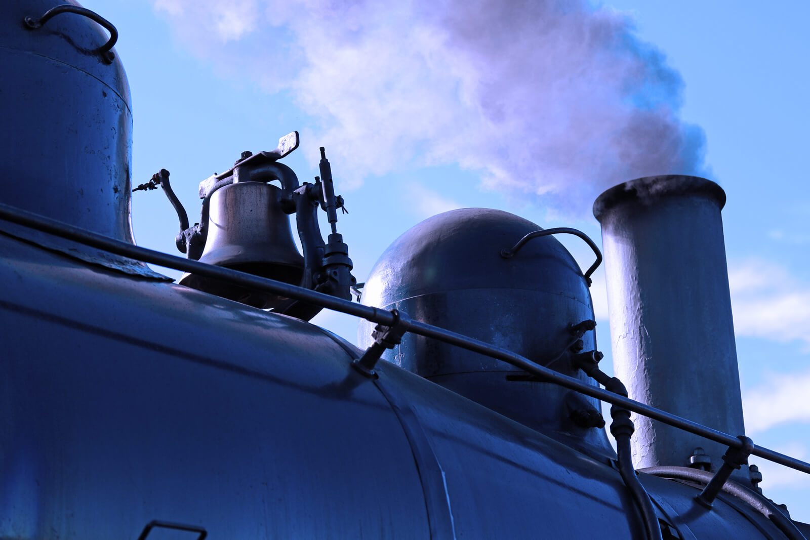 A close up of a steam engine whistle and bell with smoke coming out.